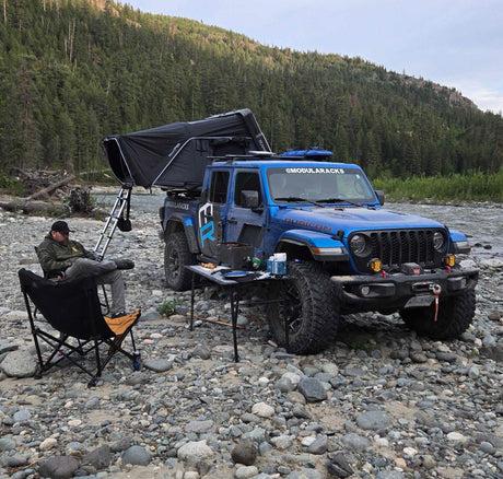 Blue Jeep Gladiator parked on a rocky riverbank with an iKamper Skycamp 4.0 Duo rooftop tent set up. A man relaxes in a camp chair next to a cooking table, with a river and forest in the background.